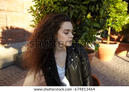 Attracractive curly female student wearing in black leather jacket sitting on the bench near orange trees in pots looking aside is enjoying the nice weather resting in city park