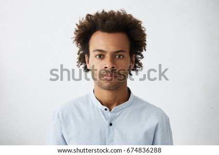 Thinking black Afro American man with serious expression looking with dark narrow eyes directly into camera while posing against white studio wall. Grave businessman in shirt thinking about work Royalty-Free Stock Photo #674836288