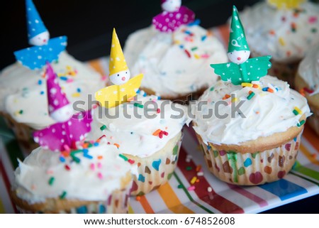 Overhead side perspective of homemade vanilla confetti sprinkle cupcakes in rainbow polka dot muffin liners topped with white frosting and colorful kids toy clown birthday cake topper in window light