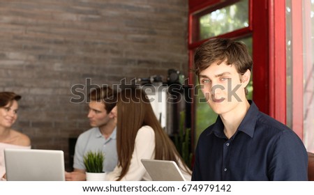 Designer in casual clothes is sitting, looking at camera and smiling, his colleagues are working in the background.