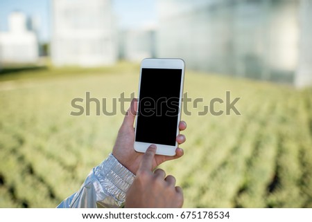 Woman holding a smart phone with blank screen on the modern hydroponic green house background
