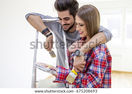 Young couple with brushes on hands looking at color palette in new flat, he's relying on ladder