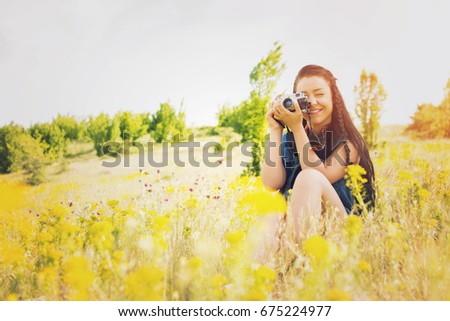 Boho female photographer outdoors in nature on sunny summer day. Cute happy young woman taking a photo in field full of yellow flowers. Matte filter, natural light.