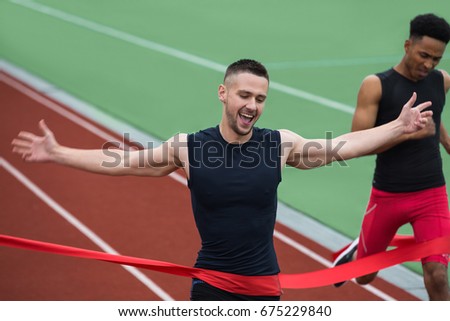 Picture of cheerful young athlete man run on running track outdoors crossing finish line. Looking aside.