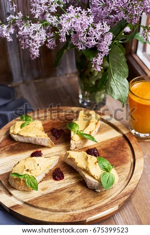 Crispy chiabatta toasts with chicken pate and onion confiture served on round wooden board on table decorated with lilac flowers. Glass of sea buckthorn berries juice. Heathy food.