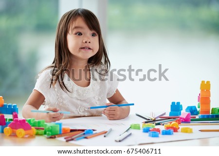 Preparing present for Mothers Day: cute little girl sitting at table in kindergarten, making funny faces and drawing colorful picture with pencils