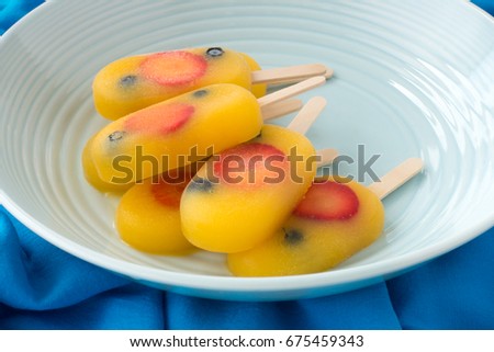 Homemade fruit ice pop, popsicle, ice cream lolly with orange juice, fresh strawberries and blueberries in a  blue porcelain bowl on dark blue fabric background. 