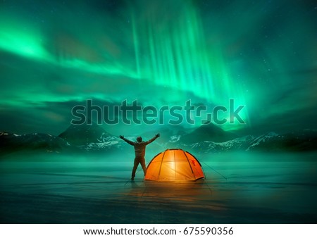 A man camping in wild northern mountains with an illuminated tent viewing a spectacular green northern lights aurora display. Photo composition. Royalty-Free Stock Photo #675590356
