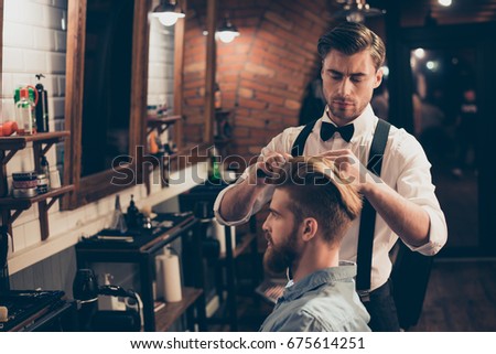 Young stylish bearded guy is in a barber shop, getting brend new haircut from a classy dressed stylist. Both are serious and attractive Royalty-Free Stock Photo #675614251