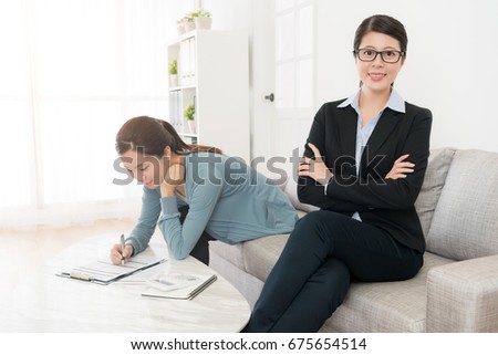 confident happy business woman sitting on sofa hands cross looking at camera smiling and her client signing insurance deal in background.