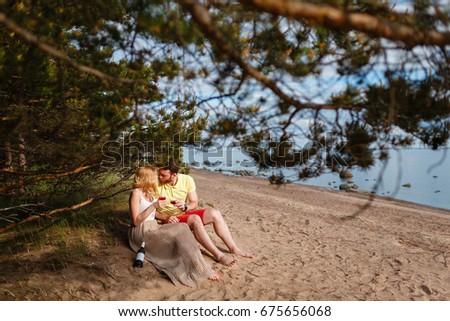 Young kissing couple sitting and drinking wine on beach on a background of stones, sea and forest, enjoy the sunset and romantic atmosphere. loving couple holding glasses and a bottle happy together