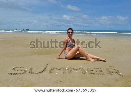 A girl in a swimsuit sits on a deserted sandy beach next to the inscription summer on the sand