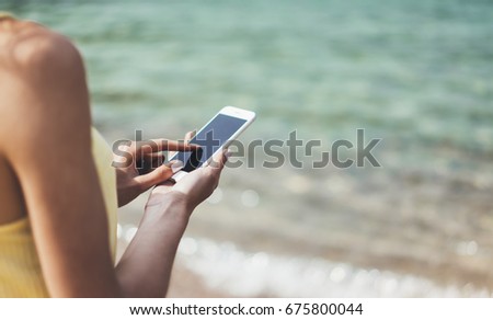 Hipster girl hold on smart phone gadget in sand coastline, mock up blank screen. Traveler using in female hand mobile on background beach seascape horizon. Tourist look on blue sun ocean, summer 