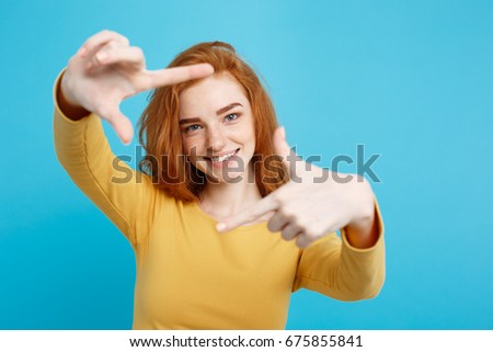 Portrait of young beautiful ginger woman with freckles cheerfuly smiling making a camera frame with fingers. Isolated on Pastel blue background. Copy space.