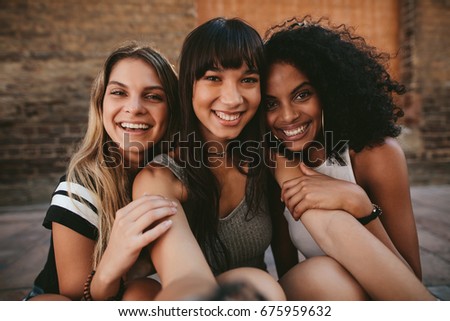 Three beautiful smiling girlfriends taking selfie with mobile phone. Multi ethnic group of women sitting outdoors by the city street and taking self portrait. Royalty-Free Stock Photo #675959632