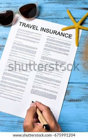 closeup of a young caucasian man signing a travel insurance contract on a colorful blue wooden table, next to a pair of sunglasses and a starfish