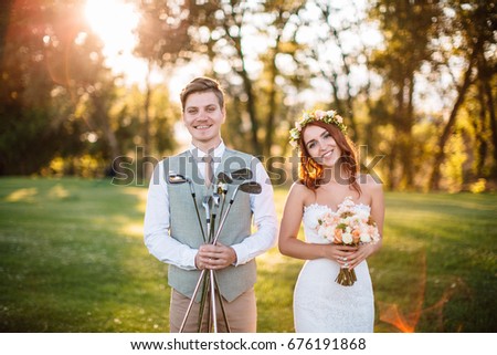 Happy smiling couple in nature. The groom is holding the golf clubs as the bride is holding a bouquet. Picture joke. Redhead bride in wreath and wedding dress. Wedding photo.