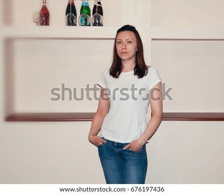 Young brunette girl in blank white t-shirt. Mockup template with a clear space for print. On the background of beige walls with shelves.