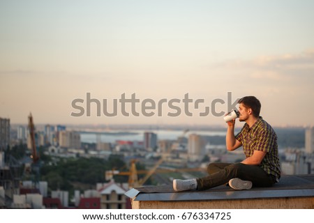 A man drinks coffee early in the morning on the roof. He looks away and thinks. In the background, the city landscape Royalty-Free Stock Photo #676334725