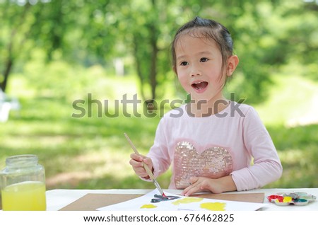 Happy asian child girl sitting at a table in a summer garden painting with paintbrush, Education art concept.