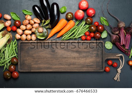 Vegetable background. A long brown wooden board, next to which lie fresh juicy organic vegetables, such as eggplants, carrots, tomatoes, asparagus beans, potatoes, beets etc, against a dark background Royalty-Free Stock Photo #676466533