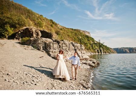 Amazing young couple holding hands on the lake shore on their sunny wedding day.