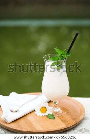 Azerbaijani and Turkish ayran - cold greek yoghurt drink - on wooden bamboo tray with straw and spoon and daisy flower in the garden by the green pool