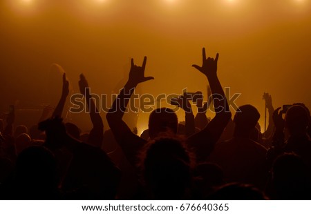 Man raising up hands with rock-n-roll gesture standing in a crowd at rock concert with musicians on the background in smoky space Royalty-Free Stock Photo #676640365
