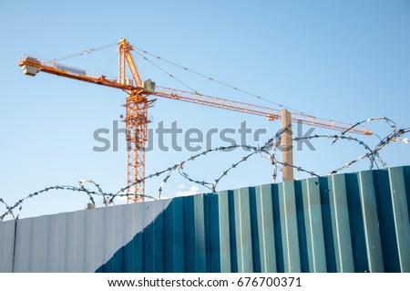 Building site with blurred crane on background and ribbed metal fence with barbwire on the foreground