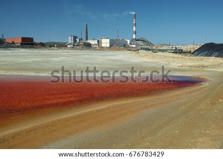 Pond with water bright red contaminated waste copper smelting production against the background of factory chimneys.