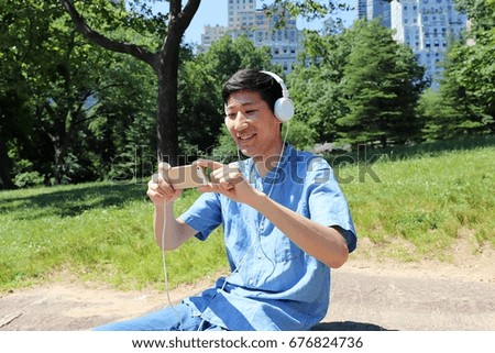 Young man sitting outside in nature park taking a picture on phone while wearing headphones 