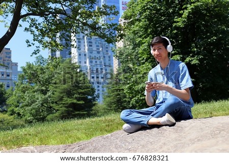 Young attractive technological man sitting in urban city park using smartphone and listening to music on white headphones 