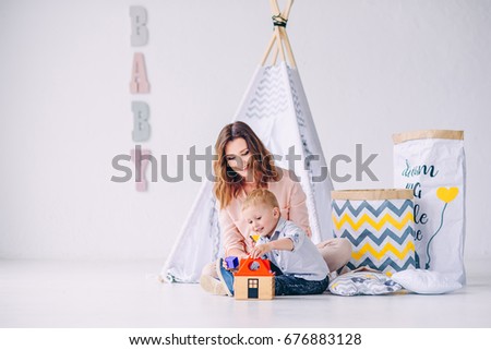 Child boy and his beautiful young mother playing together with educational toys in the light room with child's wigwam and eco-bags for toys.