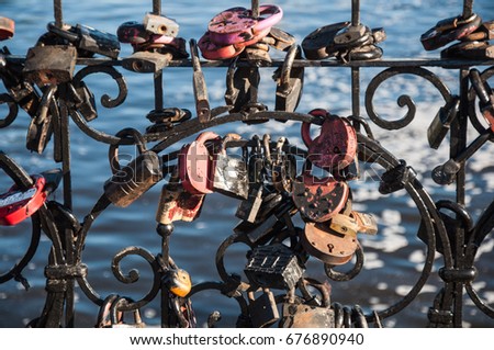 A lot of wedding padlocks of different sizes and shapes on the curved cast-iron black grate on the background of blue water