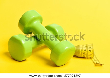 Green lightweight dumbbells and rolled tube of yellow measuring tape isolated on yellow background. Concept of slim shape and sport