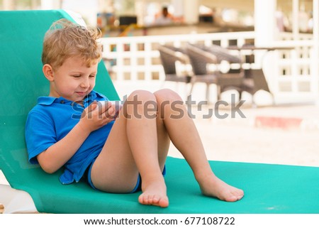 Blond cute kid is watching a cartoon or movie on a smart phone screen, holding it in his hands, sitting on a beach sunbed in a tropical resort. Bright sunny summer day.