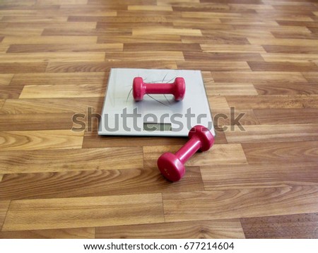 Two red dumbbells and electronic scales - the concept of excess weight. One dumbbell lies on a silver scales, and the second dumbbell lies on the brown floor near the scales