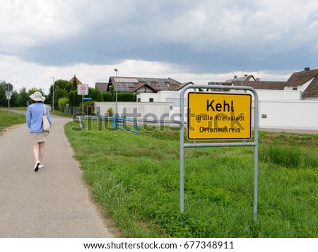 Woman walking near entrance to Kehl, Germany yellow roadsign with typical German architecture real estate buildings in the bakground and inscription Kehl, Grosse kreisstadt, Ortenaukreis , 