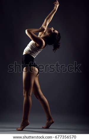 Portrait of a beautiful slender girl athlete with updo hair dancing barefoot under studio searchlights wearing black high waist panties and white top. Isolated on dark background Royalty-Free Stock Photo #677356570