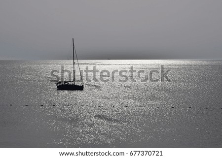 Peaceful morning, backlit black silhouette of a sailboat at tranquil sea, sunshine reflection at the metallic water surface, clear sky. Monochrome grey-colored photo