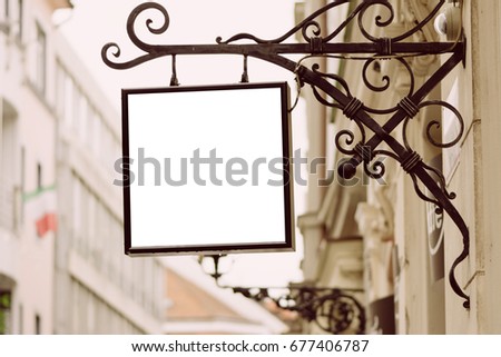 Blank square signboard, hanging from wrought iron bracket, in the city, classic architecture buildings background.