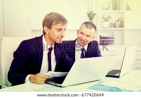 two friendly smiling young managers wearing formalwear working together using laptops in company office
