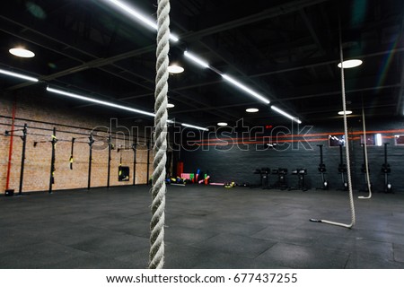 Loft big empty interior of the gym for fitness workout. Cross power training. Nobody, ropes, rowing and power rack pull-up. Stylish modern design for hipsters. Black floor and walls, red brick wall. Royalty-Free Stock Photo #677437255