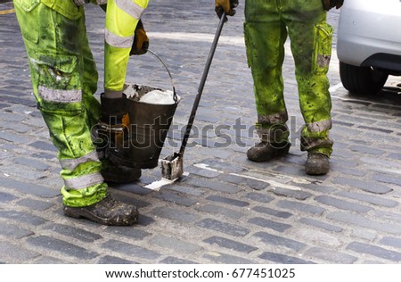 Two street maintenance workers in Hi-Vis gear and protective clothing repairing street cobbles with a bucket. Low angle shot of legs.