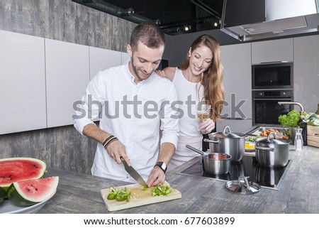 couple in the kitchen cooking vegetable soup he prepares some chili and she is drinking wine. they have a modern kitchen with grey concrete desk and glossy finish white cabinets. they wear white tops.