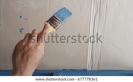 Painter's hand equipped with brush and blue color drops on carton background.