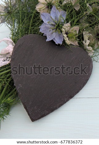 Arrangement of Nigella / Love in a Mist flowers tied with a ribbon of white lace on a pale blue wooden background with a black slate heart in front. Space for text
