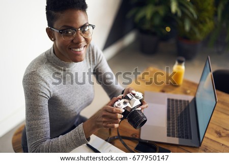 Portrait of cheerful professional afro american female freelancer earning money online editing photo made by vintage camera,happy photographer checking settings on equipment sitting in coworking space