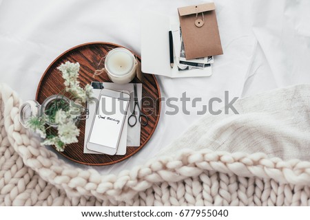 Top view of wooden tray with smartphone, old photos, candle and spring flowers on white bedding. Relaxing and posting to blog in bed at home.