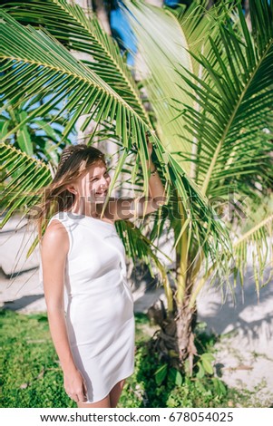 Portrait of laughing long-haired girl with amazing suntan wearing white dress near the nice green palm leave. Joyful young woman posing with smile standing near palm trees